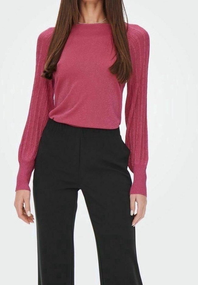 Femme portant un haut rose cintré à manches longues bouffantes et un pantalon noir taille haute à jambes larges, posant devant un fond uni.