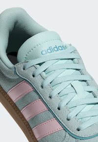 Tenisky Adidas s vrškem z mátově zelené semiše, růžovými koženými pruhy a texturovanými detaily. Mají zaoblenou špičku a gumovou podrážku.
