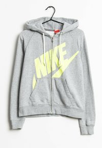 Šedá mikina na zip s kapucí a bílými šňůrkami, s velkým žlutým logem NIKE, visící na černém ramínku na bílém pozadí.