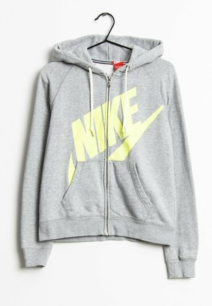 Šedá mikina na zip s kapucí a bílými šňůrkami, s velkým žlutým logem NIKE, visící na černém ramínku na bílém pozadí.