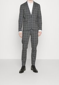 Abito grigio a scacchi realizzato in tessuto dalla texture liscia, composto da una giacca a due bottoni e pantaloni slim-fit, abbinato a scarpe nere.