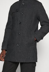 Manteau en laine gris foncé avec un motif texturé, doté d'un col montant, d'une fermeture à boutons et de poches latérales. Des boutons noirs ajoutent une touche de détail.