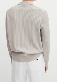 Pull en maille beige à côtes avec un col, présentant une surface texturée et des poignets ajustés, associé à un pantalon blanc.