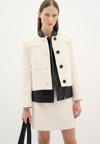 Giacca di tessuto crema con colletto e pannello in pelle nera, caratterizzata da quattro tasche frontali e bottoni neri. Indossata sopra un vestito crema abbinato.