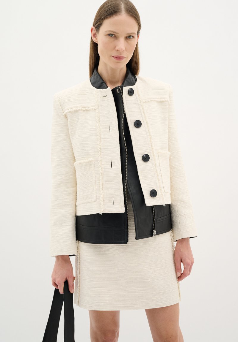 Giacca di tessuto crema con colletto e pannello in pelle nera, caratterizzata da quattro tasche frontali e bottoni neri. Indossata sopra un vestito crema abbinato.