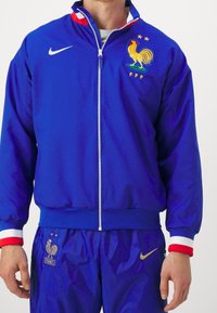 Blå Nike-jakke med frontlynlås, røde og hvide detaljer og broderet kyllingelogo med stjerner på brystet. Velegnet som sportstøj.