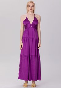 Robe maxi violette à dos nu avec un décolleté en V, design en plusieurs niveaux, détails de boutons décoratifs, fabriquée en tissu léger, accompagnée de sandales plates.