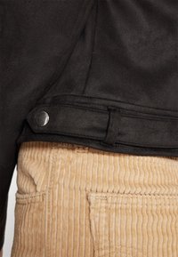 Detail van een zwarte suède jas met een zilveren drukknop, gedragen met lichtbeige corduroy broek met verticale ribbels en zakken met een patch.