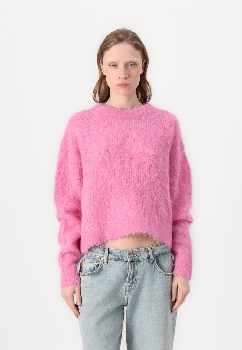 Roza skrajšan pulover iz puhastega materiala z rebrastim ovratnikom, širokimi rokavi in neenakomernim robom, v kombinaciji s svetlo modrimi jeans hlačami.