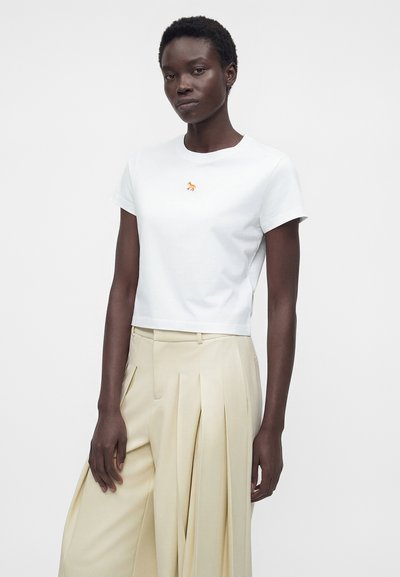 Γυναίκα που φοράει λευκό cropped t-shirt με μικρό πορτοκαλί λογότυπο αλεπούς και μπεζ παντελόνι με πιέτες, φαρδύ και ψηλόμεσο.