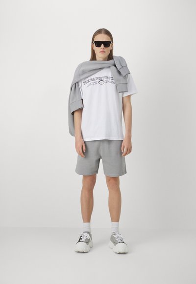 White t-shirt with graphic print, gray shorts and a gray sweatshirt draped over shoulders. Black sunglasses and chunky white sneakers.