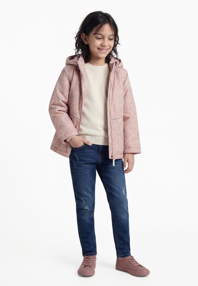 Fată zâmbitoare, purtând o jachetă cu glugă roz deschis, un pulover crem, blugi albaștri rupți și adidași roz, stând cu o mână în buzunar.