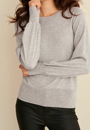Femme portant un pull gris clair tacheté à manches longues bouffantes et un pantalon en cuir noir, posant avec une main près du visage.