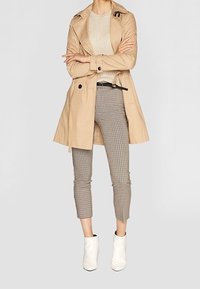 Béžový trenčkot s páskem v pase, nošený přes světlý svetr a kalhoty s krátkým střihem a vzorem psí zub; doplněný bílými kotníkovými botami.