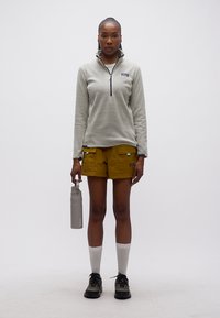 Γυναίκα όρθια, με γκρι πουλόβερ με φερμουάρ, μουσταρδί σορτς, λευκές κάλτσες, μαύρα παπούτσια, κρατώντας ένα γκρι παγούρι νερού στο πλάι της.