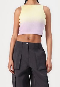 Vrouw in een mouwloze ribgebreide crop top die vervaagt van geel naar paars en een hoge taille zwarte cargobroek met grote zakken.