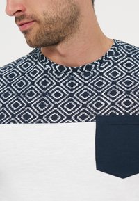 Κοντινό πλάνο ενός άνδρα που φοράει πουκάμισο με ναυτικό μπλε και λευκό μοτίβο, με μονόχρωπη ναυτικό μπλε τσέπη στο στήθος και λευκή κάτω περιοχή.