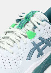Bílá sportovní bota s síťovaným svrškem, tyrkysovými akcenty a zelenými detaily. Obsahuje vzorované překryvy a perforace pro větrání.