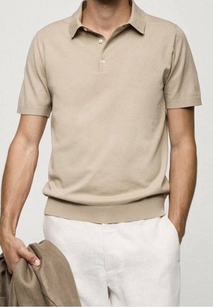 Mann trägt beige kurzärmeliges Polohemd und weiße Hose, hält eine gefaltete beige Jacke in einer Hand vor neutralem Hintergrund.