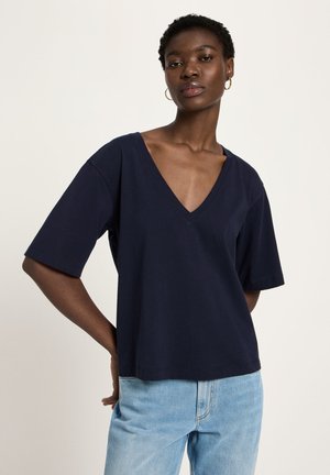 Black woman wearing a loose navy V-neck T-shirt and light blue jeans, standing with one hand behind her back against a plain background.