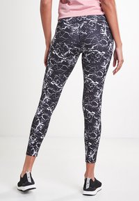 Leggings noirs avec un motif en marbre blanc. Fabriqués en matériau extensible, avec une taille haute et un design longueur cheville. Baskets noires portées.