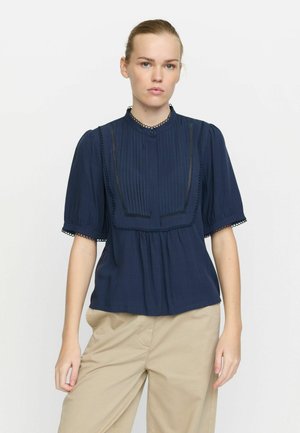 Blusa blu navy con pieghe, scollatura rotonda e maniche a sbuffo. Presenta un bordo in pizzo lungo i profili e una chiusura decorativa con bottoni.
