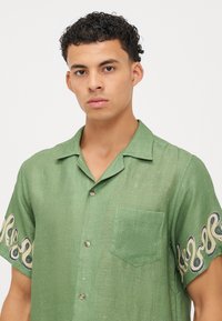 Camisa verde de manga corta en una tela texturizada. Cuenta con un bolsillo en el pecho y un distintivo bordado en espiral en las mangas cortas.