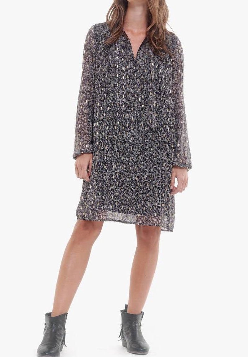 Femme portant une robe noire transparente à manches longues, longueur genou, avec des motifs blancs, et des bottines noires, debout avec une jambe légèrement avancée.