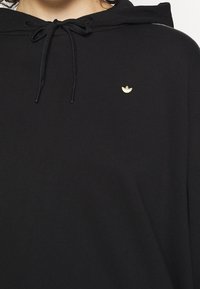 Černá mikina s kapucí, se šňůrkou a malým zlatým logem. Měkká látka, volný střih a žebrované detaily na manžetách a spodním lemu.