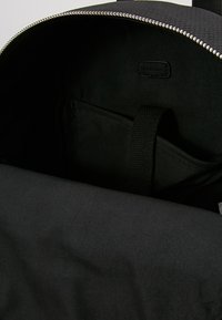 Intérieur d'un sac à dos noir montrant une ouverture zippée, une poche intérieure avec une sangle en tissu et une doublure en tissu lisse.