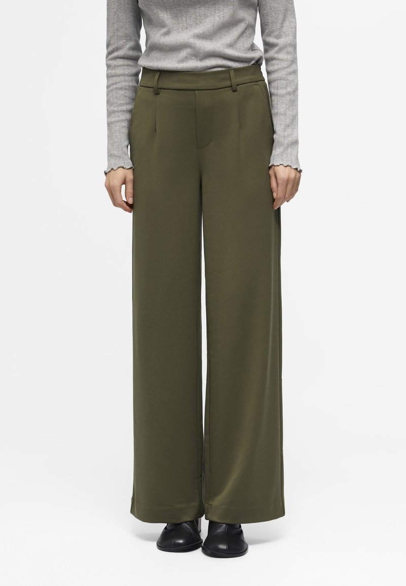 Femme portant un pantalon large taille haute vert olive avec des poches latérales, associé à un haut gris clair à manches longues et des chaussures noires.