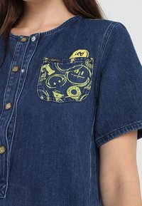 Sininen farkkupaita lyhyillä hihoilla, jossa on suuri keltakuviollinen tasku ja metallinappisuljenta edessä.