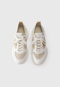 Un paio di sneakers beige e bianche con lacci, suola spessa e dettagli del logo dorati, viste dall'alto su uno sfondo bianco.