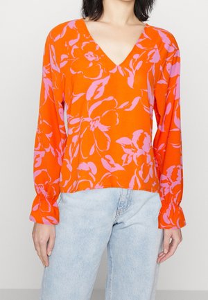Vrouw draagt een fel oranje V-hals blouse met roze bloemenpatronen en lichtblauwe spijkerbroek, staand tegen een effen achtergrond.
