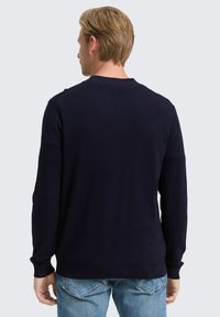 Mann mit blonden Haaren, der einen marineblauen Langarm-Pullover und hellblauen Jeans trägt und mit dem Rücken zu einem schlichten Hintergrund steht.