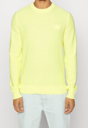 Mann trägt einen leuchtend gelben, langärmligen Strickpullover mit gerippten Bündchen und Saum, kombiniert mit hellblauen Jeans.