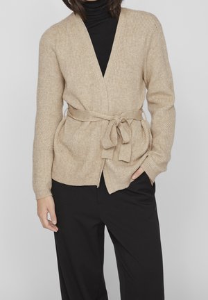 Femme portant un cardigan en maille beige avec une ceinture à la taille par-dessus un col roulé noir et un pantalon noir, debout avec une main dans la poche.