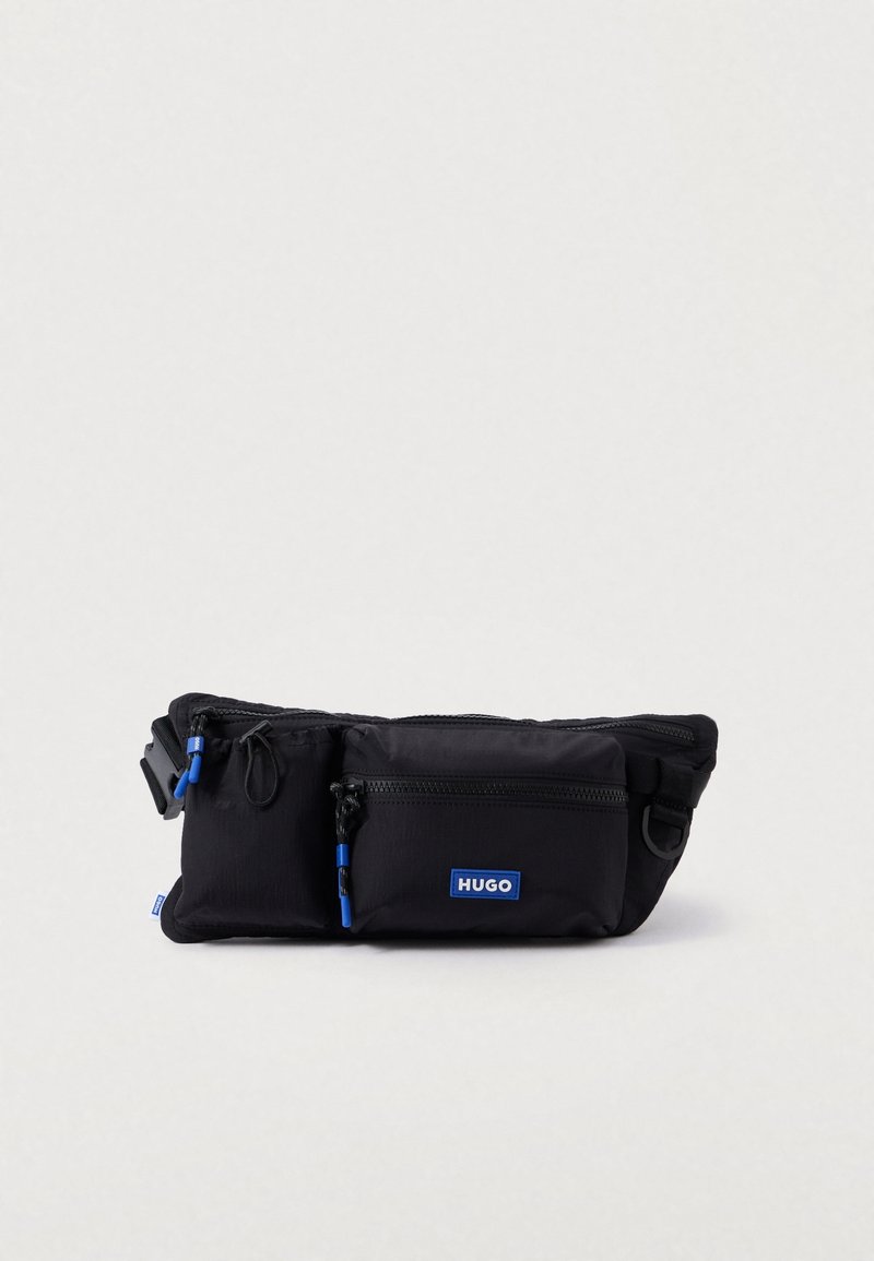 Banane noire Hugo avec deux poches zippées et un patch logo bleu, sur un fond uni clair.