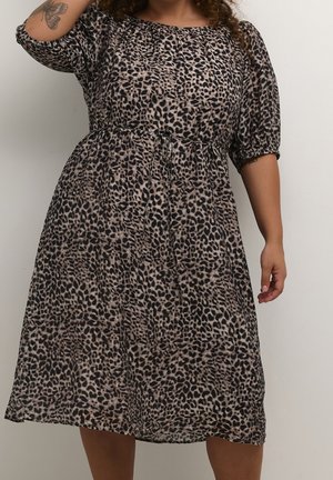 Frau trägt ein knielanges Kleid mit Leopardenmuster, Puffärmeln und einem Taillenbindegürtel, zeigt ein Schmetterlingstattoo auf ihrem linken Arm.