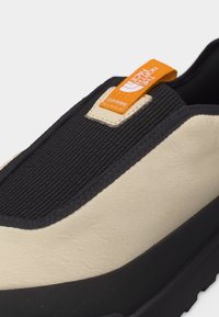 Μπεζ και μαύρο παπούτσι χωρίς κορδόνια με υφή στο μαύρο επάνω μέρος, πορτοκαλί υφασμάτινη θηλιά με ετικέτα "The North Face" και λαστιχένια σόλα.