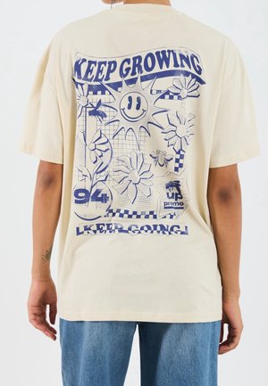 Osoba nosząca beżową koszulę z niebieską grafiką uśmiechniętego słońca, kwiatów, pszczół oraz napisami „KEEP GROWING” i „KEEP GOING” na plecach.