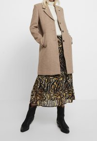 Donna che indossa un cappotto di lana beige, un dolcevita bianco, una gonna midi a fantasia con stampa animale e stivali in pelle neri fino al polpaccio, in piedi contro uno sfondo bianco.