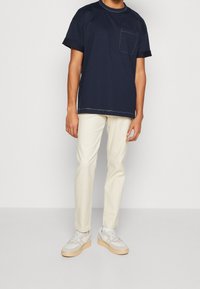 Granatowy bawełniany t-shirt z krótkimi rękawami i kieszenią na piersi, zestawiony z kremowymi zwężanymi spodniami oraz białymi tenisówkami z beżowymi podeszwami.