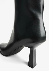 Cizma neagră din piele până la gleznă, cu vârf ascuțit și toc geometric. Textură netedă cu detalii minime pe lateral.