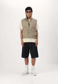 Beige padded vest with front pockets, worn over a light t-shirt. Paired with dark denim shorts and white high-top sneakers. Black sunglasses.
