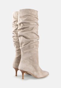 Boots bej suede până la genunchi cu detalii pliate, bot ascuțit și toc din lemn conic de 5 cm. Textură netedă cu un luciu subtil.