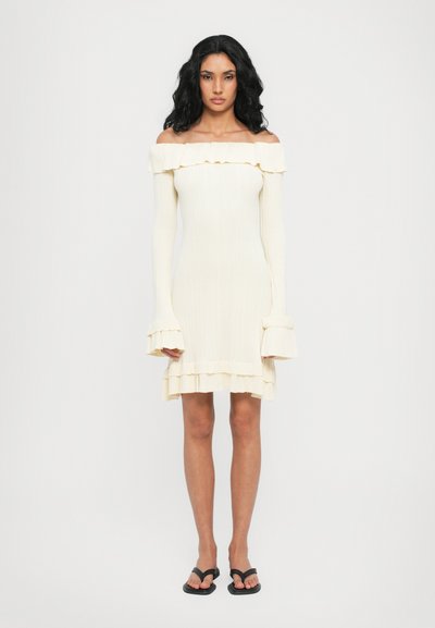 Off-the-shoulder cream ribbed dress with ruffled accents, long sleeves, and layered hem. Paired with black flip-flop sandals.