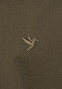 Brunt stof med et broderet kolibri-logo i en lysere beige farve, der fremhæver fine sting og en glat tekstur.