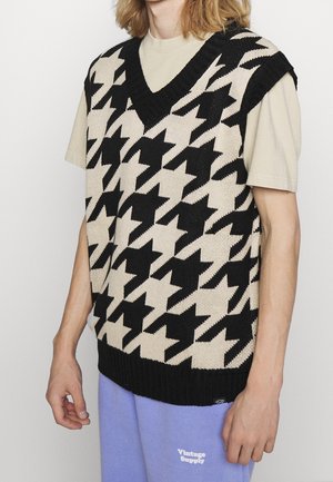 Młoda osoba nosząca czarną i beżową dzianinową kamizelkę w wzór psy ząb nad beżowym t-shirtem i jasnofioletowymi spodniami z napisem „Vintage Supply”.