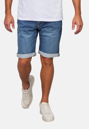 Mann trägt hochgekrempelte blaue Jeans-Shorts, weiße Sneakers und ein weißes T-Shirt und geht vor einem schlichten Hintergrund.